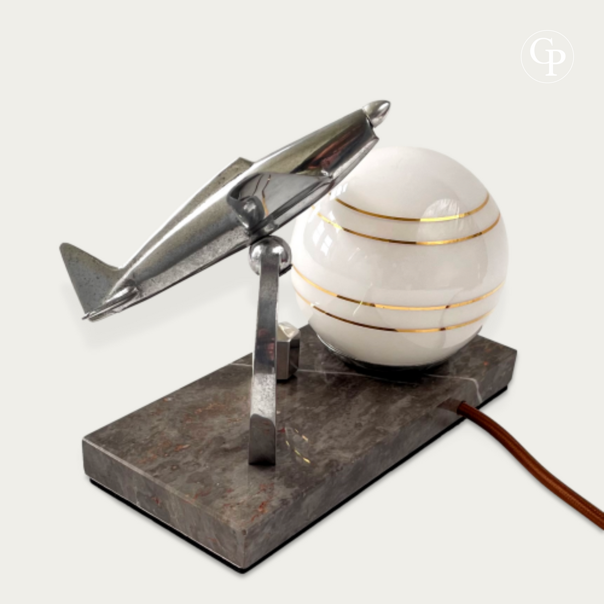 English: Vintage Art Deco desk lamp with a polished chrome Caudron Simoun airplane in flight position mounted on a marble base, next to a glowing white opaline sphere with gold stripes. Homage to Antoine de Saint-Exupéry, circa 1937.
French: Lampe de bureau Art Déco vintage avec un avion Caudron Simoun chromé en position de vol, monté sur une base en marbre à côté d’un globe opaline blanc à rayures dorées. Hommage à Antoine de Saint-Exupéry, vers 1937.