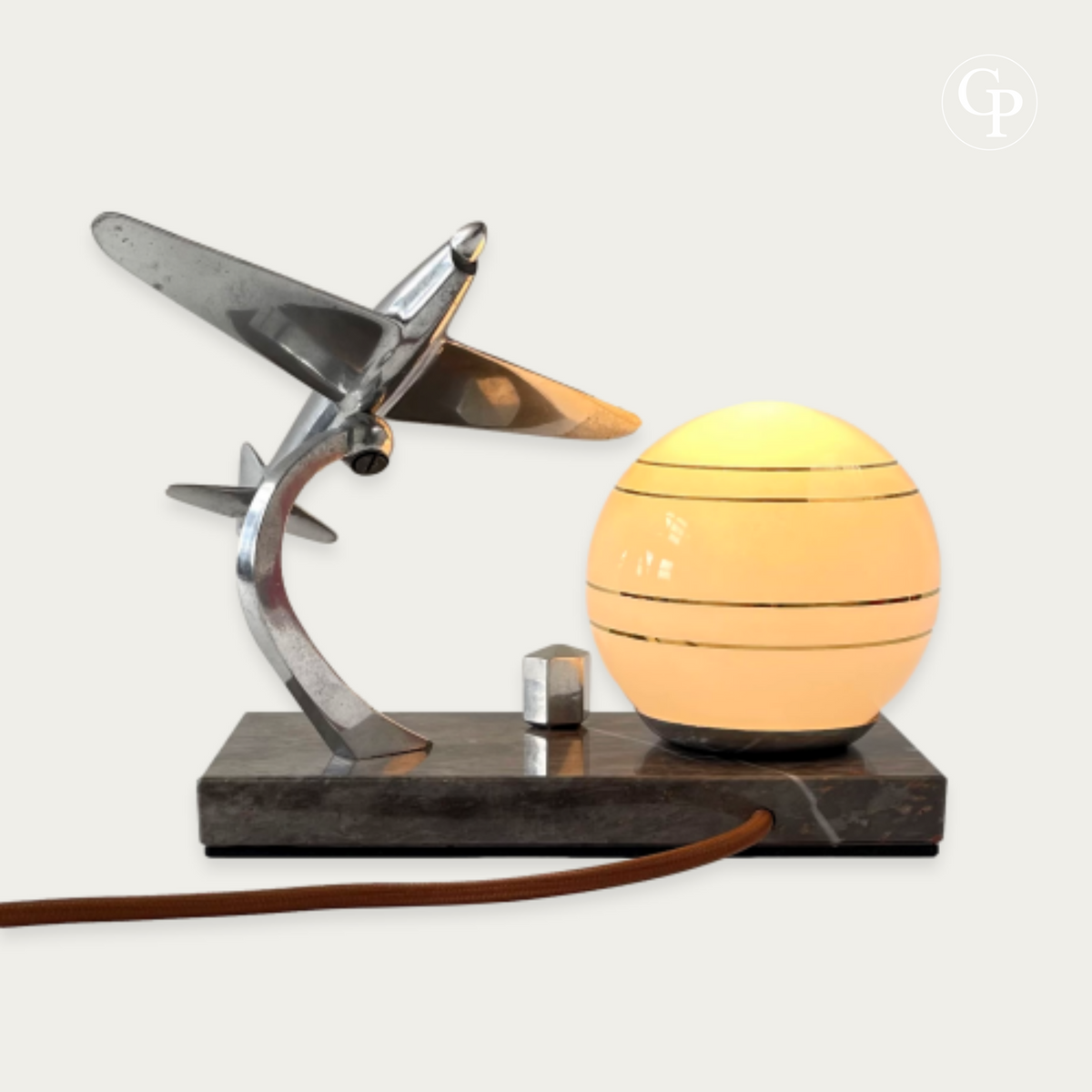 Vintage Art Deco table lamp illuminated with warm light, showing the chrome Caudron Simoun airplane in a dynamic banking position next to the glowing opaline sphere on a marble base. Homage to Antoine de Saint-Exupéry, circa 1937. | Lampe de table Art Déco vintage allumée avec une lumière chaude, présentant l’avion Caudron Simoun chromé en position de virage dynamique à côté du globe opaline lumineux sur base en marbre. Hommage à Antoine de Saint-Exupéry, vers 1937.