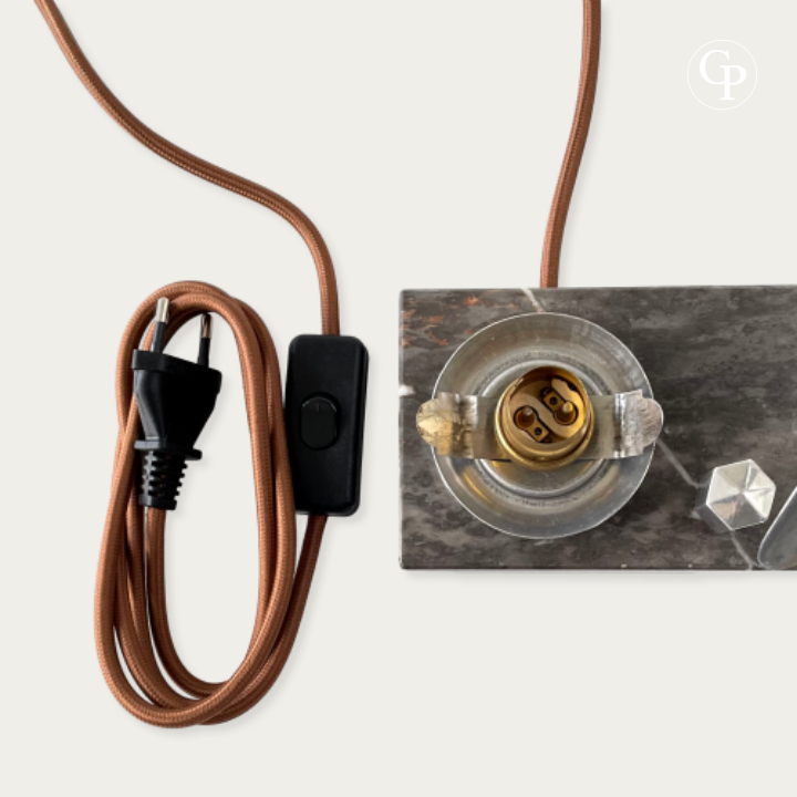 English: Close-up of the rewired and safety-compliant electrical components of the Art Deco Caudron Simoun lamp: brass bayonet socket, new brown textile cable, European plug and inline switch on the marble base. | French: Gros plan des composants électriques remis aux normes de sécurité de la lampe Art Déco Caudron Simoun : douille baïonnette laiton, nouveau câble textile brun, prise européenne et interrupteur sur la base en marbre.
