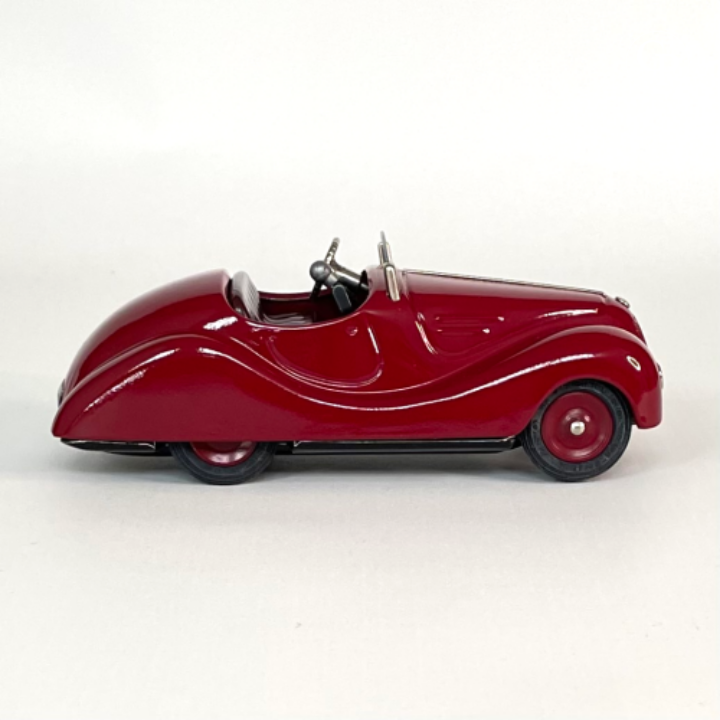 Right side profile view of the deep red Schuco Examico 4001 vintage tinplate roadster toy car, displaying its aerodynamic lines and black rubber tires on a light background. | Vue de profil droit de la voiture automate Schuco Examico 4001 de couleur bordeaux, montrant ses lignes aérodynamiques et ses pneus en caoutchouc noir sur fond clair.